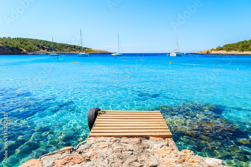 Wooden jetty and view of azure blue sea with sailing boats in distance, Cala Portinatx bay, Ibiza island, Spain