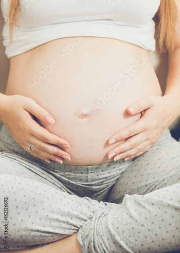 Young pregnant woman sitting on couch and holding hands on tummy