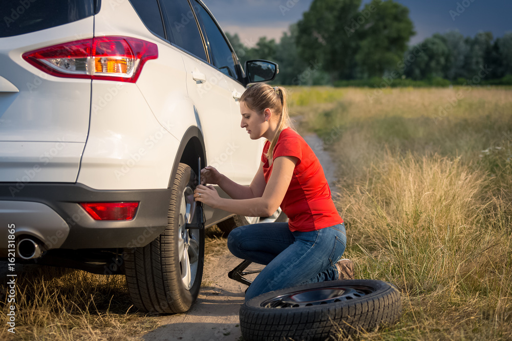 Young woman changing car wheel on the rural road going through field Young woman changing car wheel on the rural road going through field