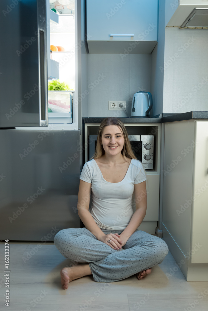 Smiling woman in pajamas sitting on floor at kitchen next to open refrigerator at evening