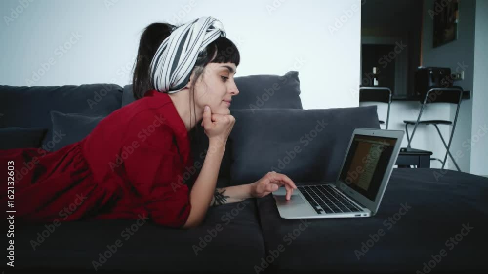 Young pretty brunette lady in red dress with white striped turban lies on sofa at home and looks on monitor of laptop, texting comments for shared photos in social network and smiles. Young pretty brunette lady in red dress with white striped turban lies on sofa at home and looks on monitor of laptop, texting comments for shared photos in social network and smiles.