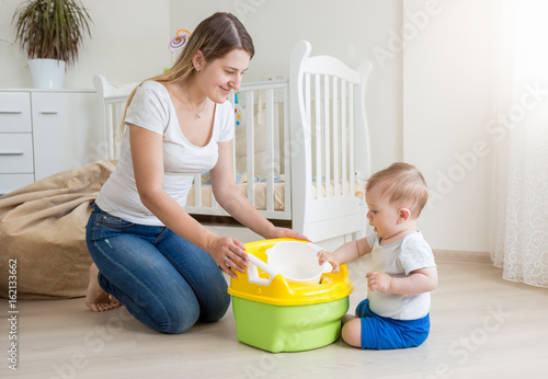 Young smiling mother sitting on floor at living room and teaching her baby boy how to use chamber pot photo