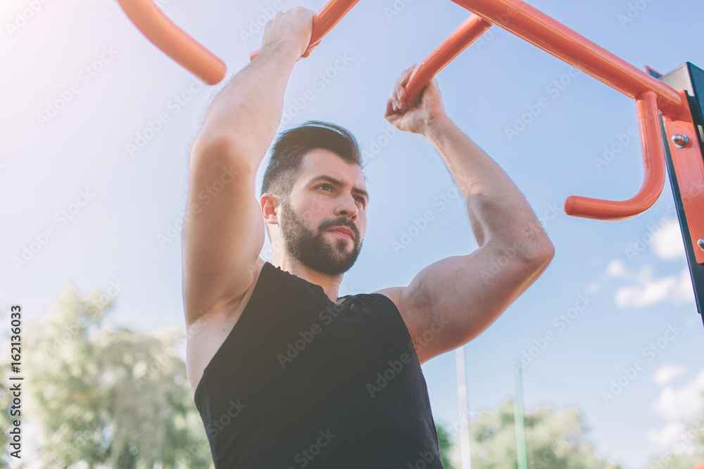 Closeup of strong athlete doing pull-up on horizontal bar.Mans fitness with blue sky in the background and open space around him. Young man with sportive clothes in the city Closeup of strong athlete doing pull-up on horizontal bar.Mans fitness with blue sky in the background and open space around him. Young man with sportive clothes in the city