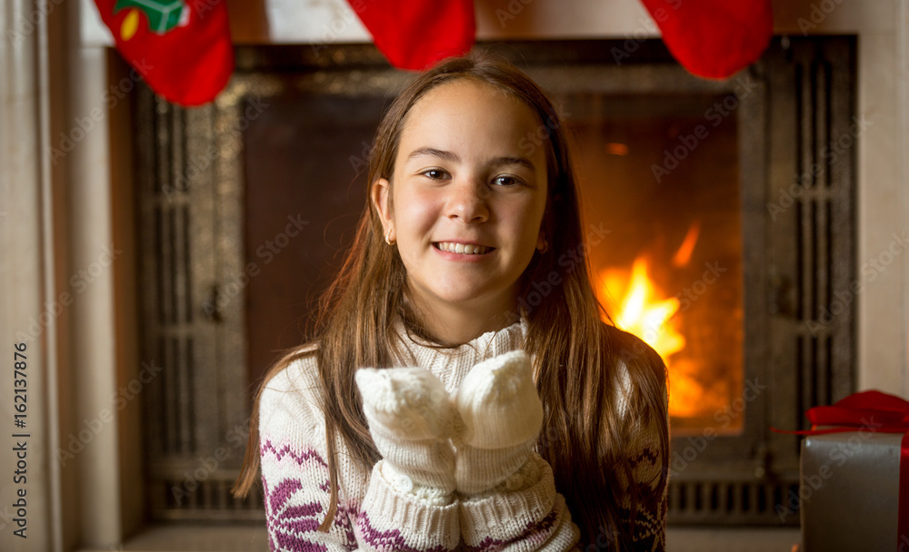 Happy smiling girl in sweater and gloves posing at burning fireplace and decorated Christmas tree Happy smiling girl in sweater and gloves posing at burning fireplace and decorated Christmas tree