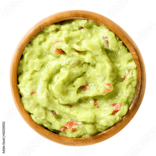 Guacamole in wooden bowl. Also short guac, a light green dip or salad, made of mashed avocados, tomatoes, onions, garlic, lemon, cayenne pepper and salt. Macro food photo close up on white background. photo