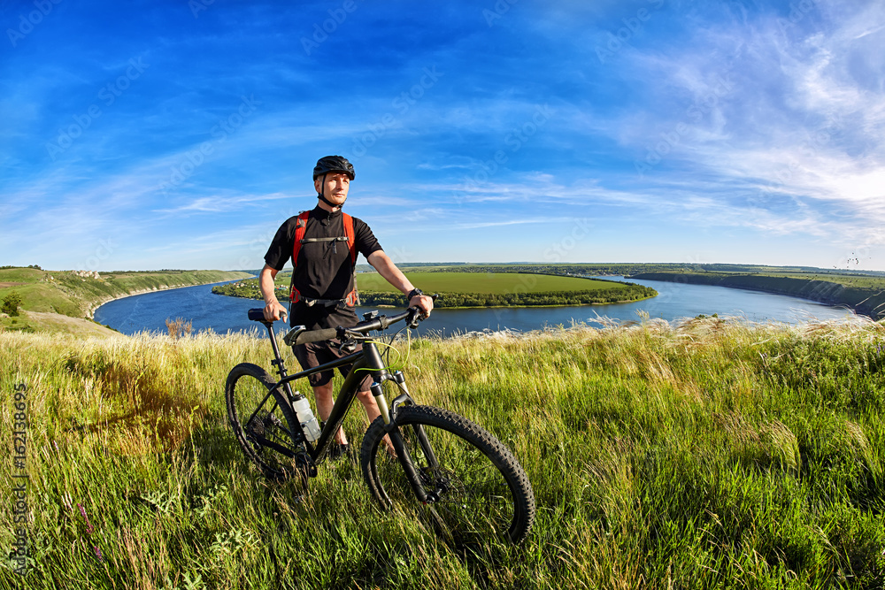 Attractive mountain cyclist stands on the hill above the beautiful river. Attractive mountain cyclist stands on the hill above the beautiful river.