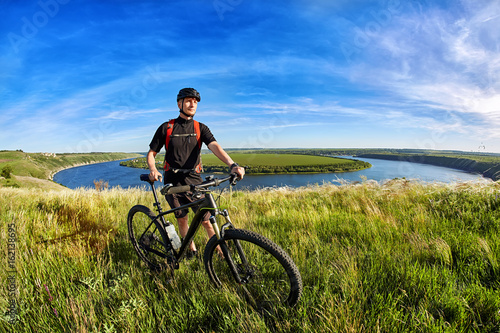 Attractive mountain cyclist stands on the hill above the beautiful river.