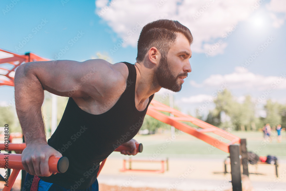 Obraz premium A portrait of a focused muscular bearded man in black workout clothes doing dips on parallel bars . Mans fitness with blue sky in the background and open space around him. Sports and crossfit