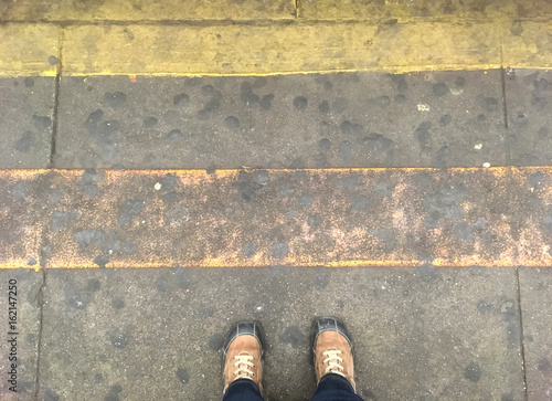 Standing in a New York subway station platform looking down at feet next to the yellow safety line, grime all over the concrete floor of the platform.  Gross, disgusting, and filthy subway station.