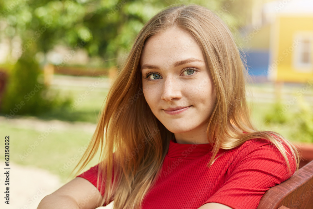 Fototapeta premium Headshot of beautiful blonde girl having warm eyes, freckles and sincere smile looking pleased into camera while having rest at bench in garden. People, facial expressions and lifestyle concept