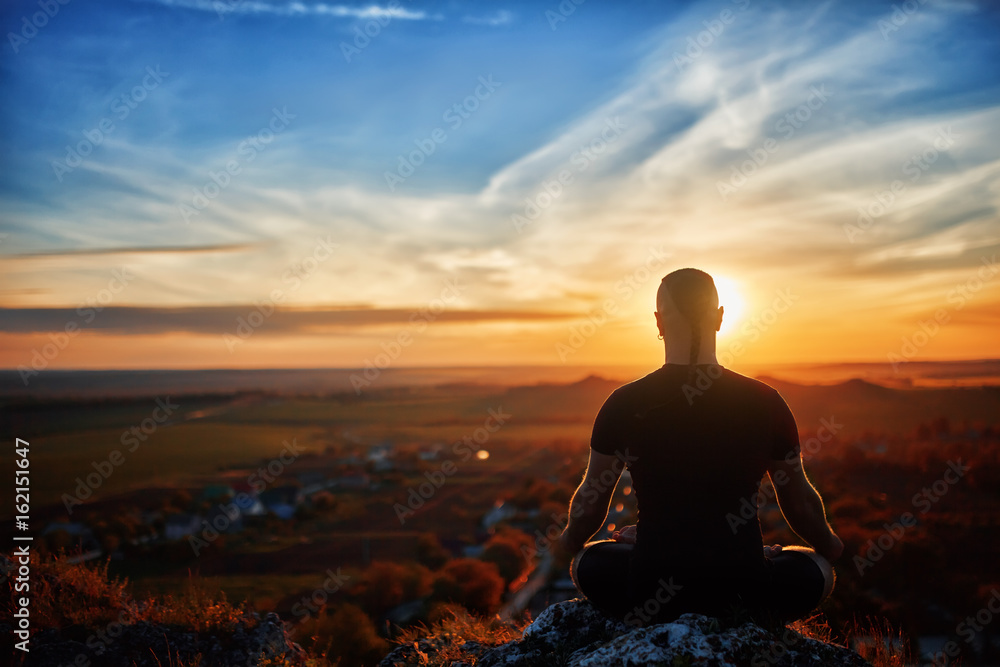 Rear view of the man meditating yoga in lotus pose on the rock at sunset. Rear view of the man meditating yoga in lotus pose on the rock at sunset.