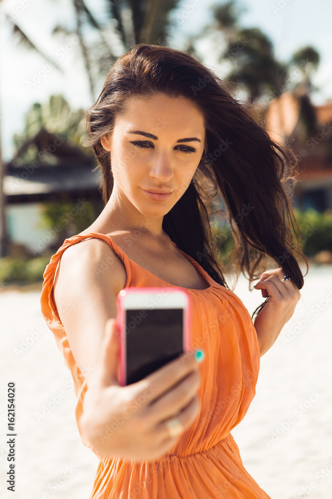 Close up of sexy stylish hipster woman in light dress, sunglasses make  selfie self portrait on a smartphone digital photocamera on a tropical  beach. Swag urban teen girl. Outdoor lifestyle portrait. Stock