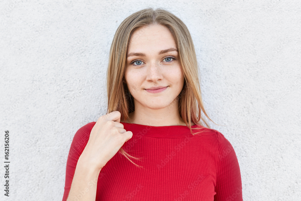Foto Attractive young female with fair hair, blue eyes and freckles posing against white background touching her hair with hand dressed in red clothes