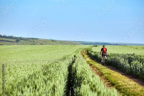 Rear view of the young cyclist with backpack cycling in the track of the field. © Aleksey