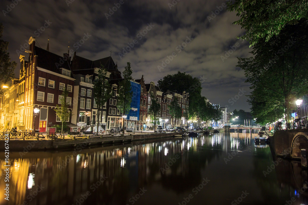 Beautiful traditional old buildings at night along the canal with reflection on water in Amsterdam, the Netherlands