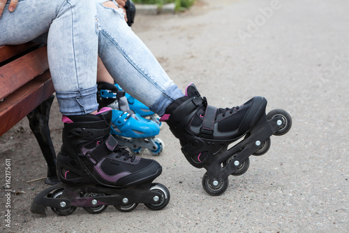Human feet are wearing in-line skates. Woman and kid sitting on the bench while rollerblading