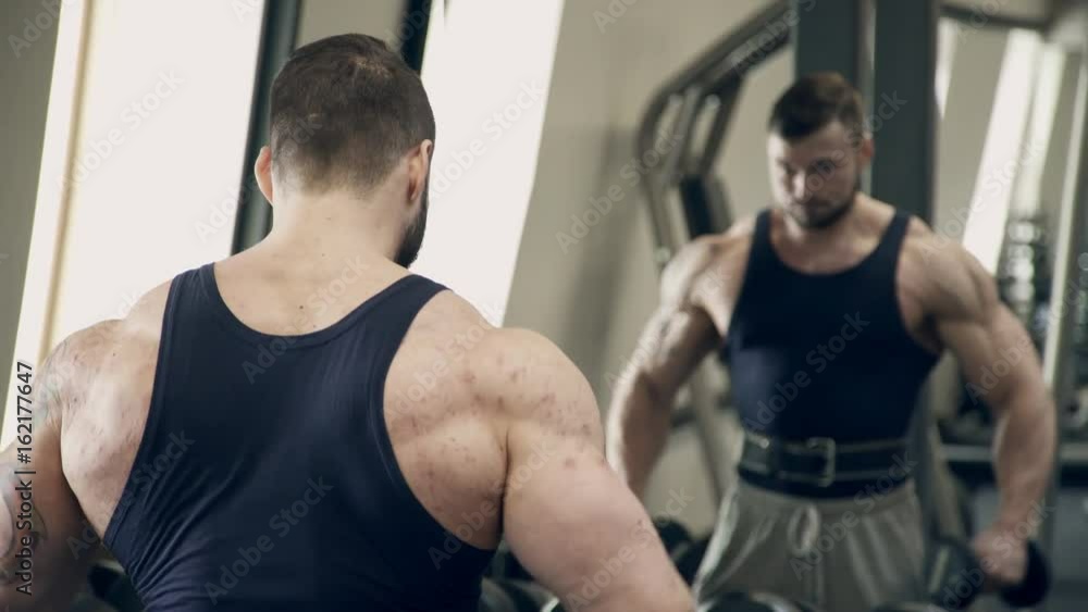 Man athlete doing dumbbell lateral raises in gym indoors. Standing directly in front of mirror with exhale he lifts black dumbbells up to level of shoulders, hands smoothly move through sides in plane Man athlete doing dumbbell lateral raises in gym indoors. Standing directly in front of mirror with exhale he lifts black dumbbells up to level of shoulders, hands smoothly move through sides in plane