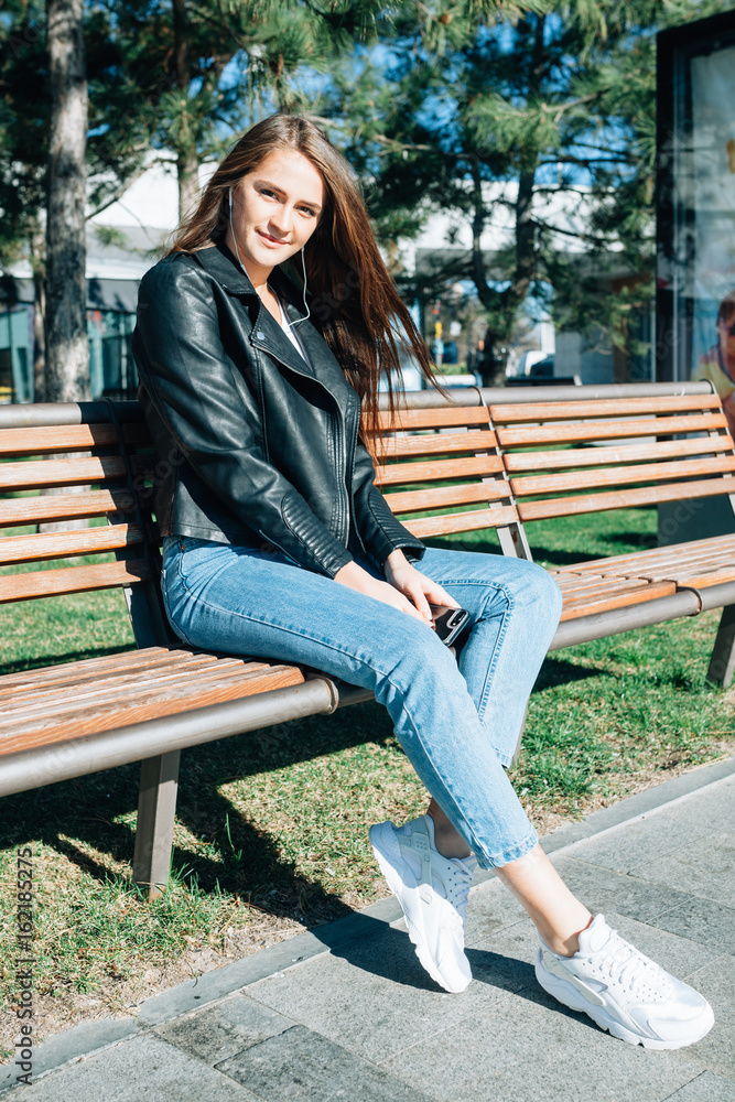 The lovely girl sits on a bench in the park in one of sunny days. She listens to music in earphones and gently smiles. On her light jeans, white sneakers and a black leather jacket. The lovely girl sits on a bench in the park in one of sunny days. She listens to music in earphones and gently smiles. On her light jeans, white sneakers and a black leather jacket.