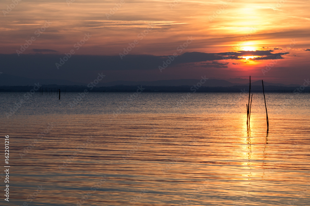 Beautiful sunset with sun behind some clouds and reflecting on a lake, with red and orange tones in the sky and clouds Beautiful sunset with sun behind some clouds and reflecting on a lake, with red and orange tones in the sky and clouds