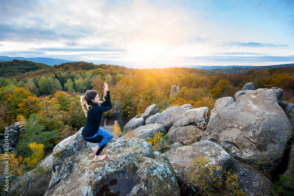 Attractive young female is practicing yoga and doing asana Garudasuna on the top of the huge boulder in the evening. Beautiful sunset, autumn forests, rocks and hills on the background