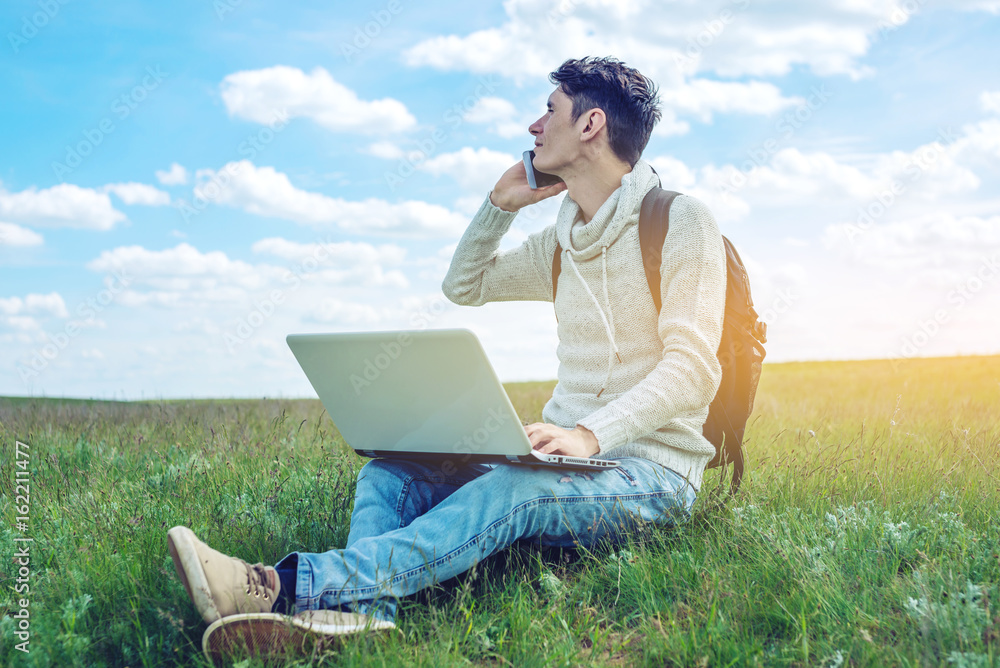 Young man sitting on a green meadow with laptop and talking on the phone on the background of blue cloudy sky Young man sitting on a green meadow with laptop and talking on the phone on the background of blue cloudy sky
