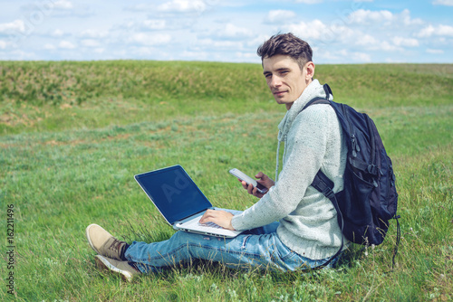 Young man sitting on a green meadow with laptop and using the phone on the background of blue cloudy sky