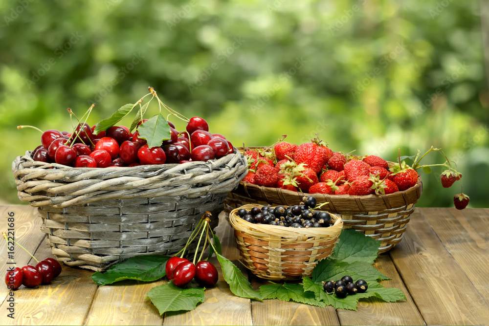 Summer gifts: sweet cherry, strawberry and black currant in wicker baskets on wooden desk on background of green leaves Summer gifts: sweet cherry, strawberry and black currant in wicker baskets on wooden desk on background of green leaves