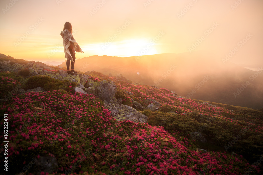 Obraz premium young woman hiking at mountain peak above clouds and fog. Hiker girl wrapping in warm poncho outdoor. Young girl over the clouds. Wonderful landscape with cloud inversion. Flowers in the mountains