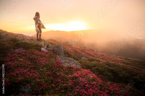 young woman hiking at mountain peak above clouds and fog. Hiker girl wrapping in warm poncho outdoor. Young girl over the clouds. Wonderful landscape with cloud inversion. Flowers in the mountains