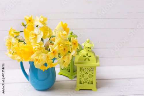 Fototapeta Naklejka Na Ścianę i Meble -  Spring daffodils or narcissus flowers in  blue pitcher and decorative green lanterns on white  wooden background.