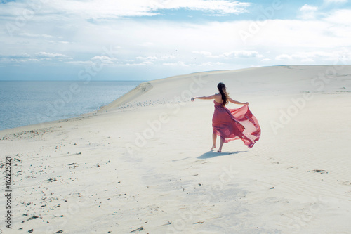Woman in red waving dress with flying fabric runs on background of dunes. Enjoy the freedom