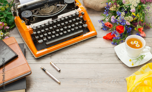 Orange vintage typewriter, books and a bouquet of summer field flowers on a gray background.