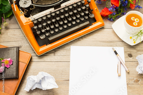 Workspace with orange vintage typewriter with white blank. Bouquet of summer field flowers, pen and cup of chamomile tea.