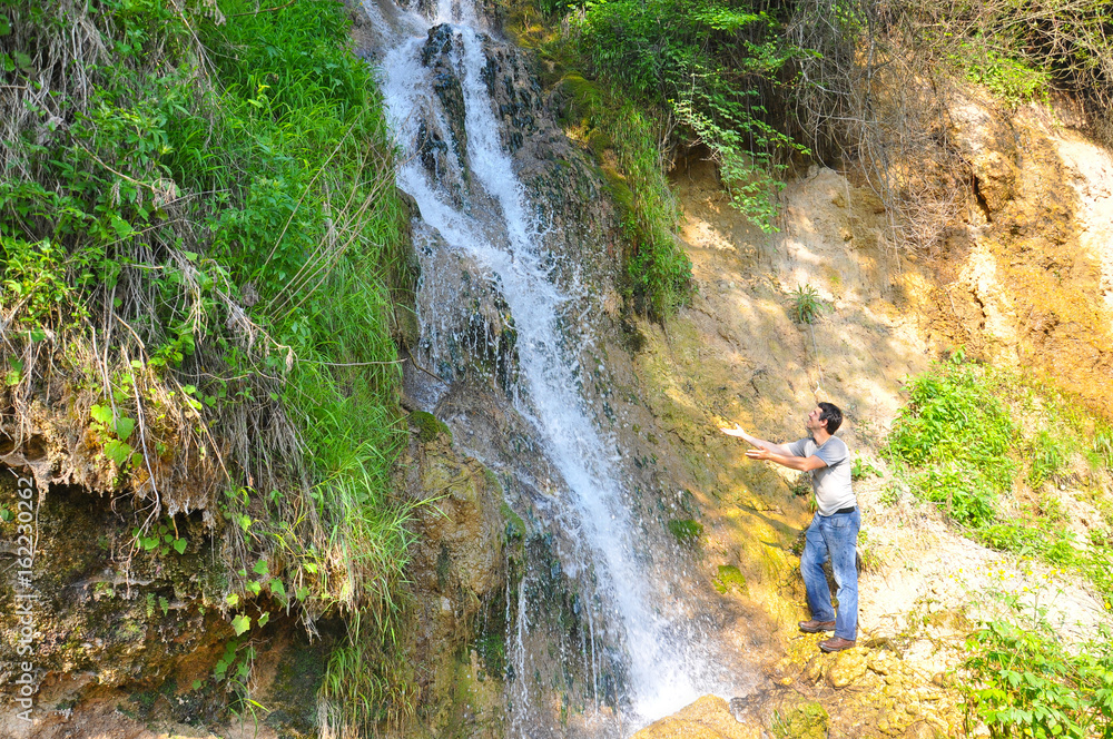 Man under a waterfall on morning sun. Happy hiker under a small waterfall in mountains. Man in wild nature.