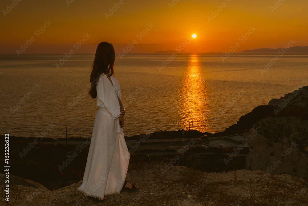 Young girl on the edge of a cliff in a white dress watching the sunset in Santorini, Greece. Female in white sundress on summer Europe travel vacation. Young girl on the edge of a cliff in a white dress watching the sunset in Santorini, Greece. Female in white sundress on summer Europe travel vacation.