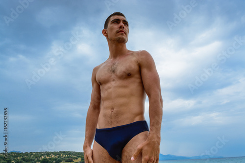 Muscular man standing on the beach in a speedo. The concept of freedom, power, sport, healthy lifestyle.