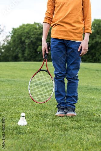 Cropped shot of little boy holding badminton racquet and standing on green grass with shuttlecock near by