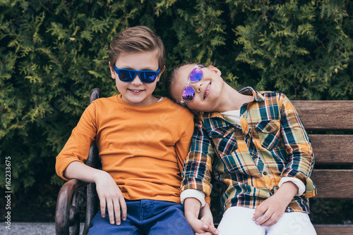 smiling children in sunglasses holding hands while sitting on bench at park