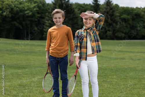 Adorable little children holding badminton racquets and smiling at camera outdoors