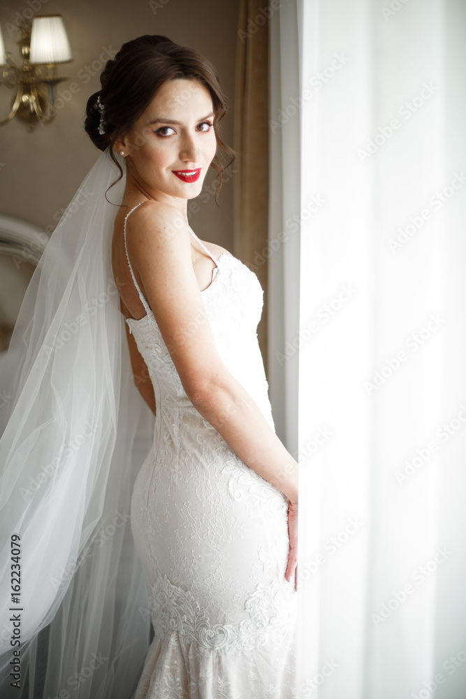 Bride in rich wedding gown stands before a window in the room Bride in rich wedding gown stands before a window in the room