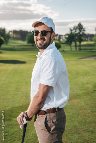 Handsome man in sunglasses holding golf club and smiling at camera