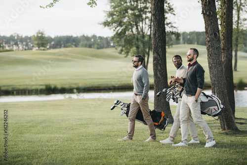 Multiethnic golf players holding bags with golf clubs and walking on golf course photo