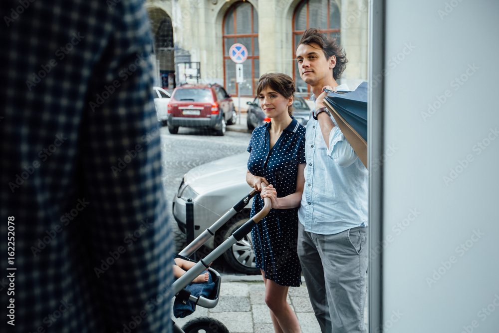 Photo taken through the window. Shopping, parenthood and lifestyle concept. Couple with paperbag and baby carriage choosing a suit or shirt in the mens classic suit shop Photo taken through the window. Shopping, parenthood and lifestyle concept. Couple with paperbag and baby carriage choosing a suit or shirt in the mens classic suit shop
