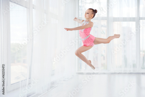 Fototapeta Naklejka Na Ścianę i Meble -  Little gymnast girl in pink sportswear dress, performing art gymnastics element, jumping, doing split leap in the air