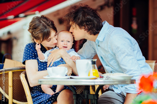 Happy parenthood: young parents kissing and calming down their sweet baby boy in street cafe.