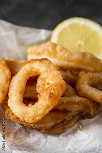 Seafood. Squid rings fried in mesle with lemon and ketchup on a ceramic plate. Dark background. Snack to beer.