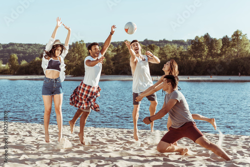 young smiling friends playing beach volleyball on riverside at daytime