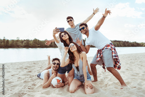 group of cheerful friends taking selfie together on smartphone on beach