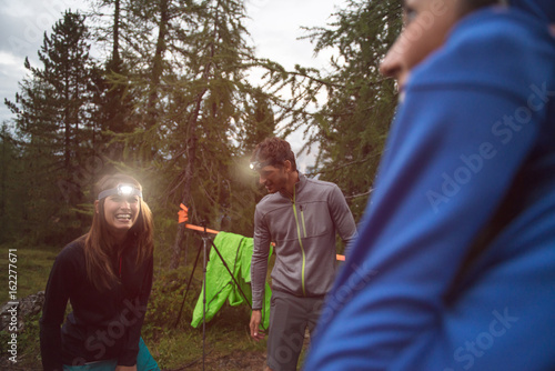Happy smiling woman and man with headlamp flashlight during evening near camping. Group of friends people summer adventure journey in mountain nature outdoors. Travel exploring Alps, Dolomites, Italy. photo