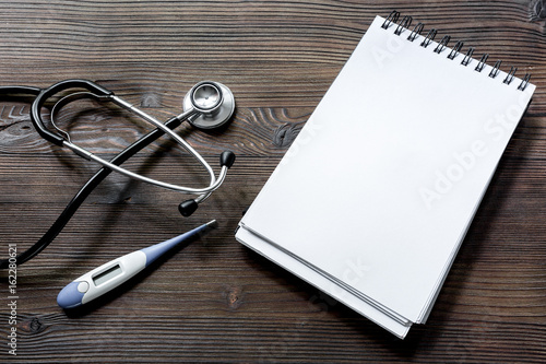 phonendoscope, thermometer and notebook on dark wooden desk top view make an appointment with doctor mock up